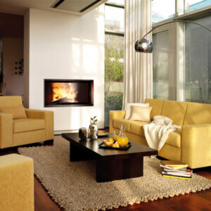 A modern, built-in fireplace insert Lina 120 H/S featuring a large black-framed square glass door and bright, active flames. The fireplace is set into a smooth white mantelpiece with a light stone base. A blue sofa with a grey herringbone blanket, a stack of white books, and a window showing a snowy mountain landscape define the contemporary living room. Cork, Homexpert