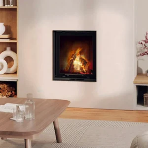A square, black-framed fireplace insert, LINA 67 S, showing a vibrant wood fire, built into a minimalist white wall. The contemporary room features light wooden flooring, a light wood coffee table with glassware, and built-in wooden shelving visible on the left side. Cork, Homexpert