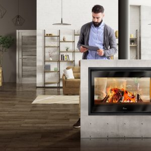 Double Sided Stove: Nickel. Modern black double-sided wood-burning stove on a pedestal base, featuring a large glass window, with a smiling couple admiring the fire in a bright, open-plan living room and kitchen area, Cork, Homexpert