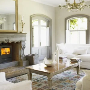 Bright, elegant living room with large windows, white sofas, a wooden coffee table, and a classic stone fireplace featuring a Country 575 modern wood-burning stove with visible flames. Cork, Homexpert