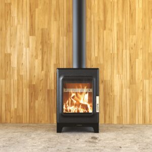 Vertical black THEODORE steel wood-burning stove with a large glass door showing bright flames, installed against a natural wooden wall with a matching black flue and concrete floor, Cork, Homexpert